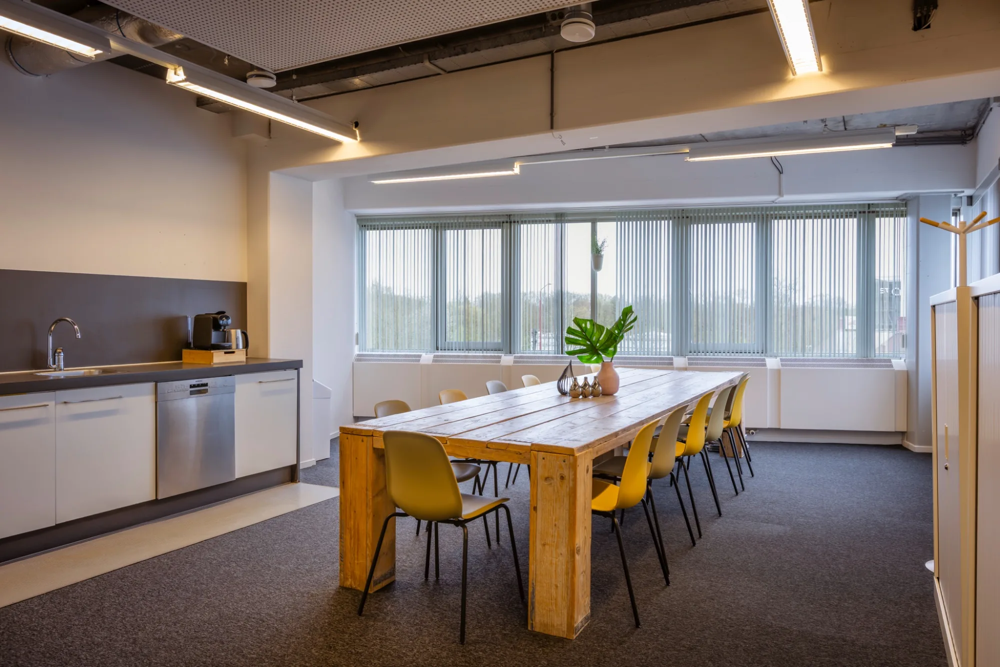 Ruime lunchkamer met grote houten tafel, gele stoelen en daglicht via lamellengordijnen
