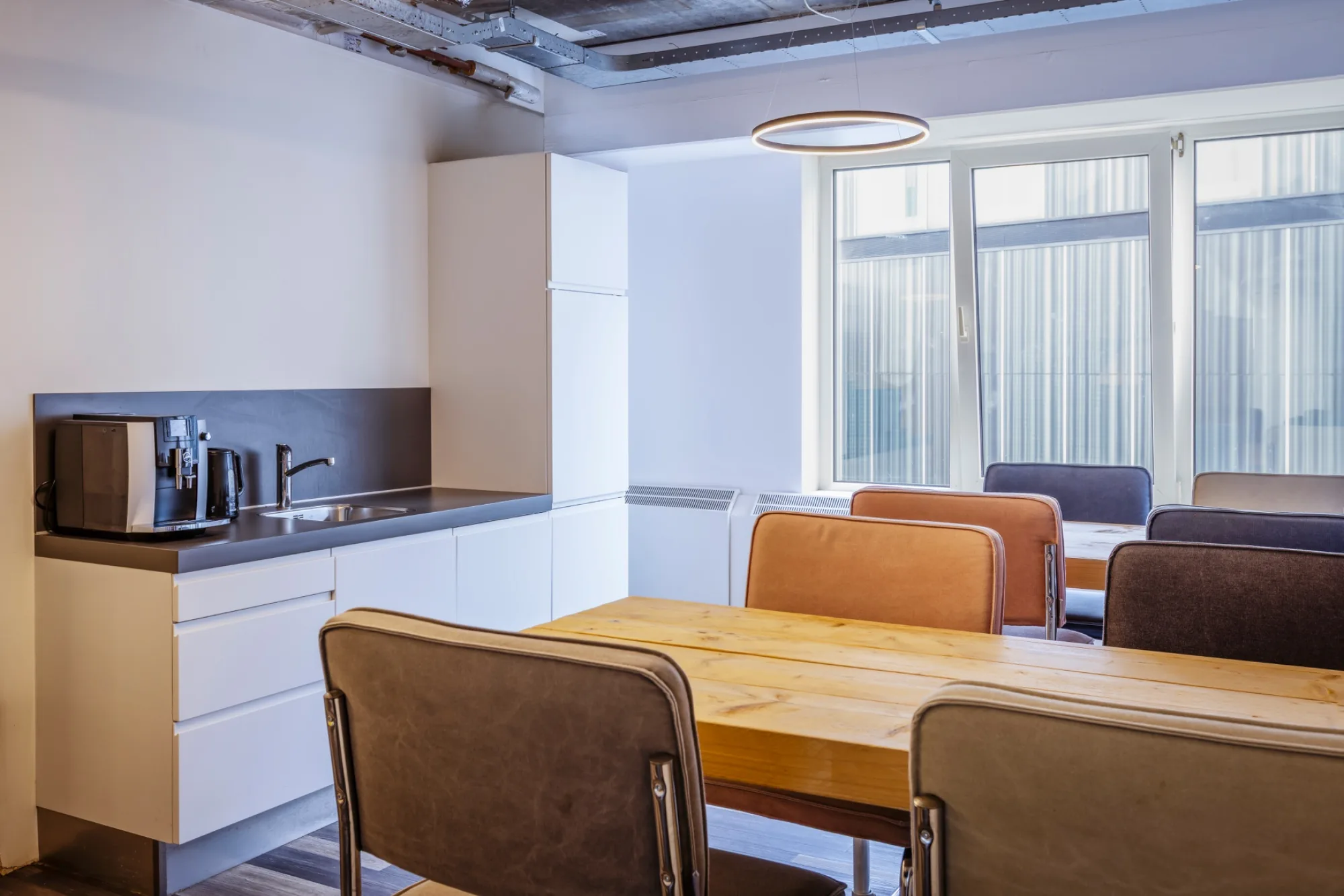 Lichte kantoorpantry met witte keukenopstelling, houten tafel, leren stoelen en ronde hanglamp